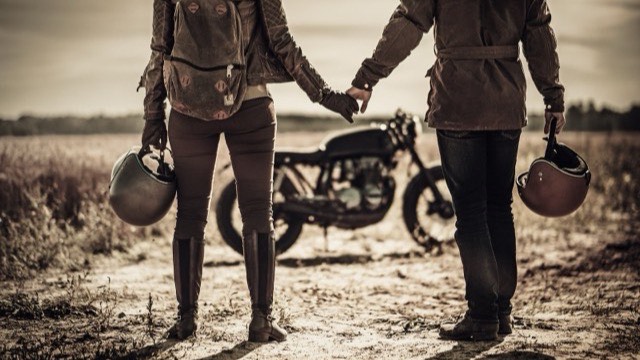 A couple holding hands and carrying their helmets beside a cafe racer motorcycle in a sepia-toned field