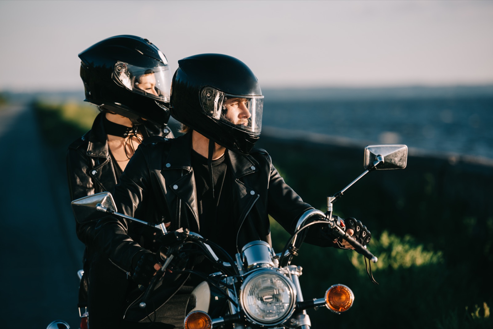 A rider and pillion passenger in helmets with visors up, sitting on a motorcycle by the coast