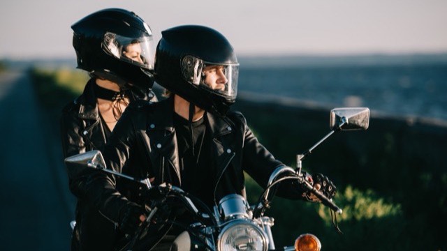 A rider and pillion passenger in helmets with visors up, sitting on a motorcycle by the coast