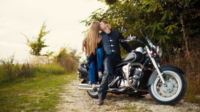 A couple kissing while leaning against a cruiser motorcycle on a tree-lined dirt path