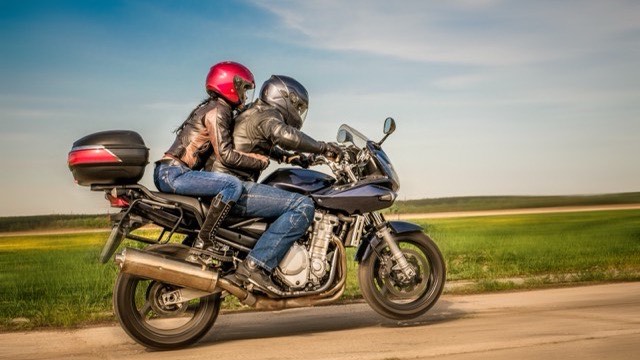 A rider and pillion in helmets riding two-up along an open country road on a sunny day